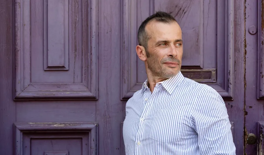 Photo portrait d'André debout devant une porte violette. André a été diagnostiqué avec une SEP récurrente-rémittente et a souffert de problèmes urinaires et intestinaux. 