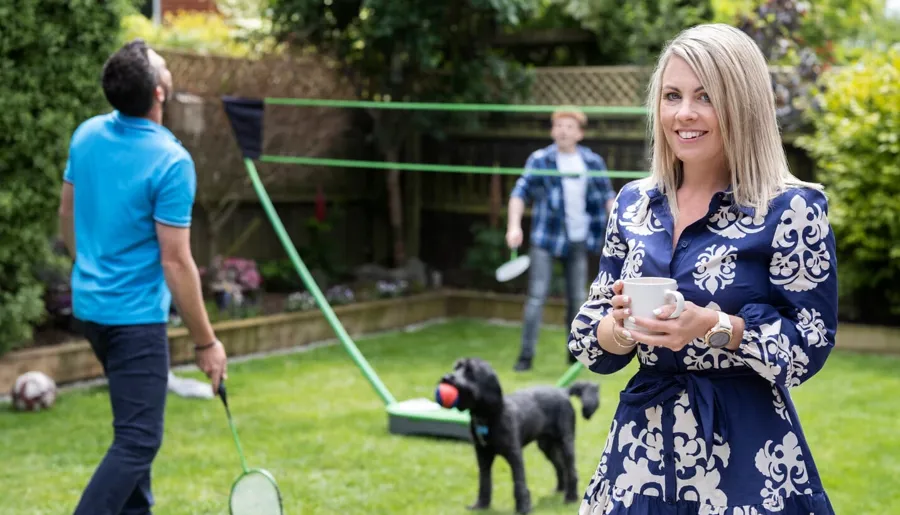 Laura se tient dans son jardin avec sa famille qui joue au badminton en arrière-plan. Laura a trouvé une solution pour l’aider à gérer ses problèmes intestinaux résultant de sa SEP.